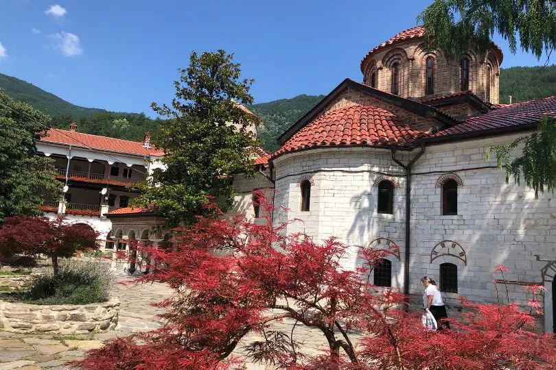 Das Bachkovo Kloster in Bulgarien mit seinen weißen Wänden und roten Dächern. Dahinter ragen bewaldete Berge auf.