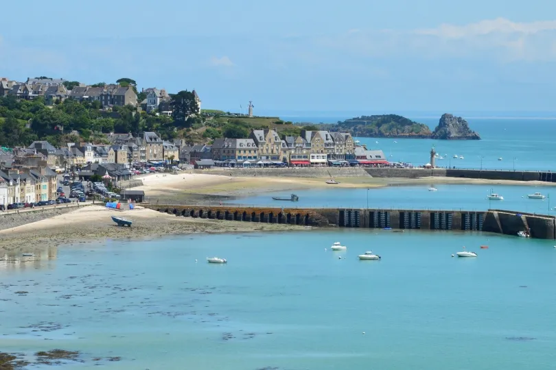 Küstenort in der Bretagne mit Hafen, Sandstrand und traditionellen Steinhäusern. Mehrere kleine Boote liegen im türkisblauen Wasser, während eine lange Hafenmauer die Bucht begrenzt.