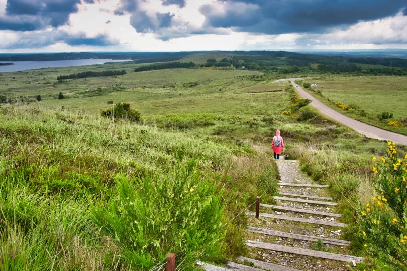Panoramic view of the Monts d’Arrée in Brittany, with rolling green moorland, a winding country road, a lake in the distance, and a hiker descending a hillside path beneath dramatic clouds.