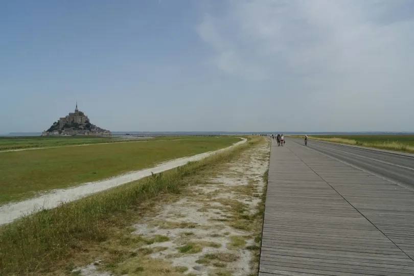 Blick über die weite Küstenlandschaft auf den Mont-Saint-Michel in der Ferne. Ein langer Weg führt durch flaches Marschland zur markanten Klosterinsel, während einige Menschen zu Fuß und mit dem Fahrrad unterwegs sind.