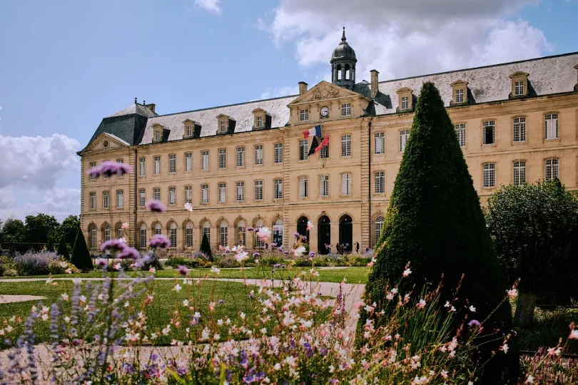 Historisches Klostergebäude mit französischen Flaggen, umgeben von einem gepflegten Garten mit Blumenbeeten und kegelförmig geschnittenen Bäumen.