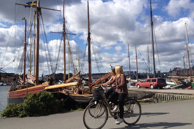 Zwei Frauen fahren mit ihren Fahrrädern durch Stockholm, vorbei an den Segelbooten im Hafen.