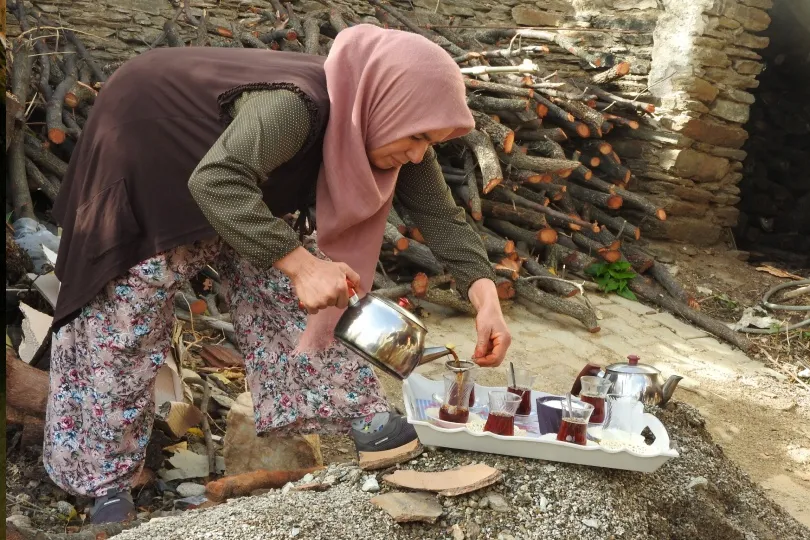 Eine Frau mit Kopftuch schenkt im Freien türkischen Tee in mehrere Gläser ein. Im Hintergrund liegen gestapelte Holzscheite vor einer alten Steinmauer.