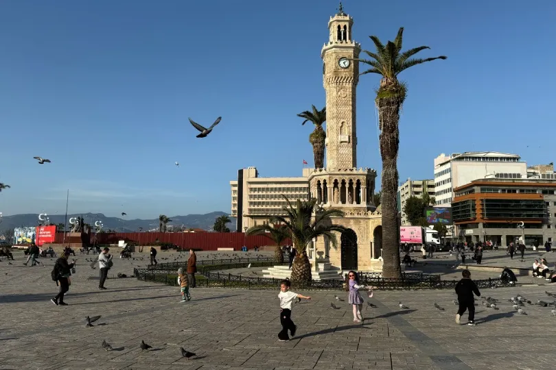 Ein belebter Platz in Izmir mit dem historischen Uhrturm, hohen Palmen und zahlreichen Tauben. Menschen spazieren über den Platz, während Kinder zwischen den Vögeln spielen.