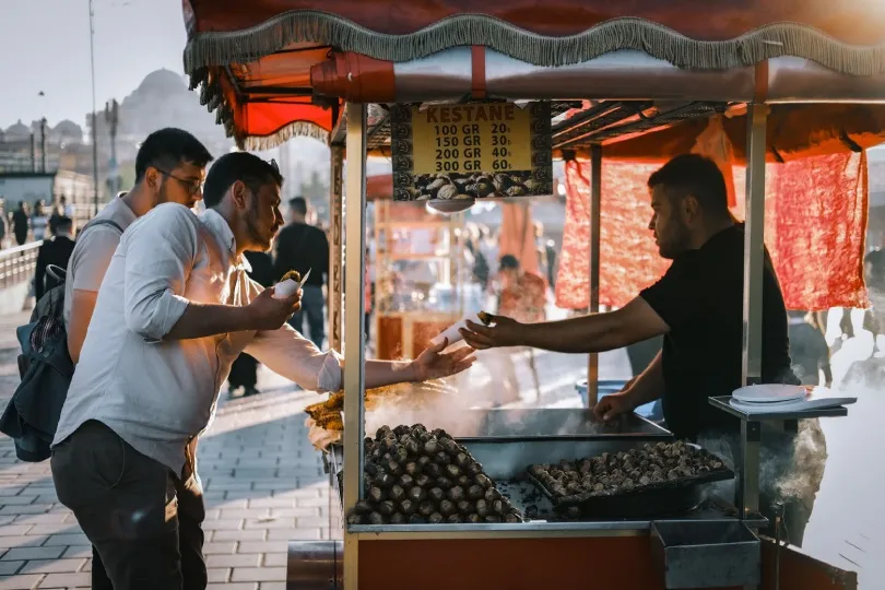 Ein Straßenverkäufer verkauft frisch geröstete Kastanien an einem Stand, während zwei Männer davor warten und probieren.