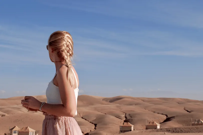 Eine blonde Frau mit geflochtenem Zopf, weißem Top und rosafarbenem Rock steht in der Agafay-Wüste bei Marrakesch in Marokko. Hinter ihr erstreckt sich eine weite, hügelige Steinwüstenlandschaft mit vereinzelten Camp-Zelten unter einem leicht bewölkten blauen Himmel.