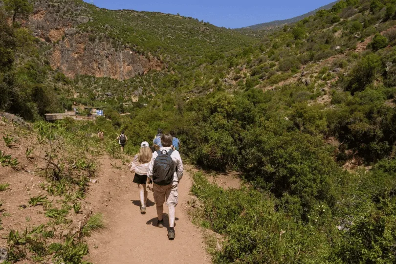 Eine Gruppe von Wanderern mit Rucksäcken folgt einem schmalen, staubigen Wanderweg durch die grüne Berglandschaft von Akchour in Marokko. Dichte Sträucher, Bäume und felsige Berghänge umgeben den Weg unter einem klaren blauen Himmel.