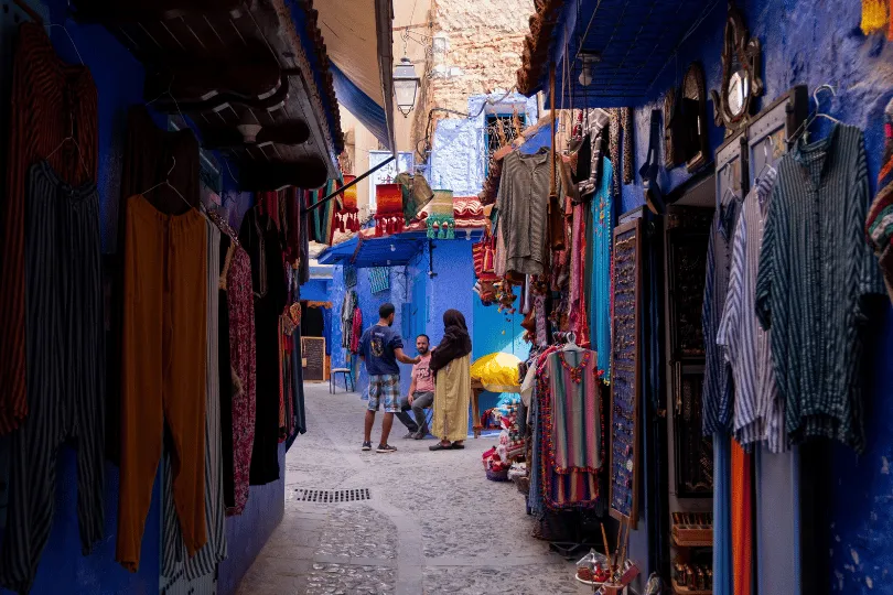 Schmale Marktgasse in der Medina von Chefchaouen, Marokko. Zwischen den leuchtend blau gestrichenen Häusern hängen bunte Kleidungsstücke, traditionelle Gewänder und Souvenirs vor kleinen Geschäften. Mehrere Menschen unterhalten sich in der gepflasterten Gasse, während Sonnenlicht auf die blauen Fassaden fällt.