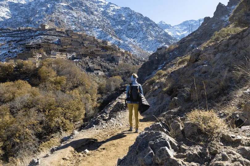 Ein Wanderer mit Rucksack steht auf einem schmalen, felsigen Bergpfad bei Imlil im Hohen Atlas in Marokko. Am gegenüberliegenden Berghang liegt ein traditionelles Berberdorf mit dicht aneinandergereihten Steinhäusern. Im Hintergrund erheben sich mächtige, schneebedeckte Berggipfel unter einem klaren blauen Himmel.