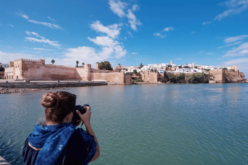 Eine Frau fotografiert vom Ufer aus eine historische, von Mauern umgebene Küstenstadt. Im Hintergrund stehen helle Gebäude und eine Festungsanlage unter blauem Himmel, im Vordergrund liegt ruhiges Wasser.
