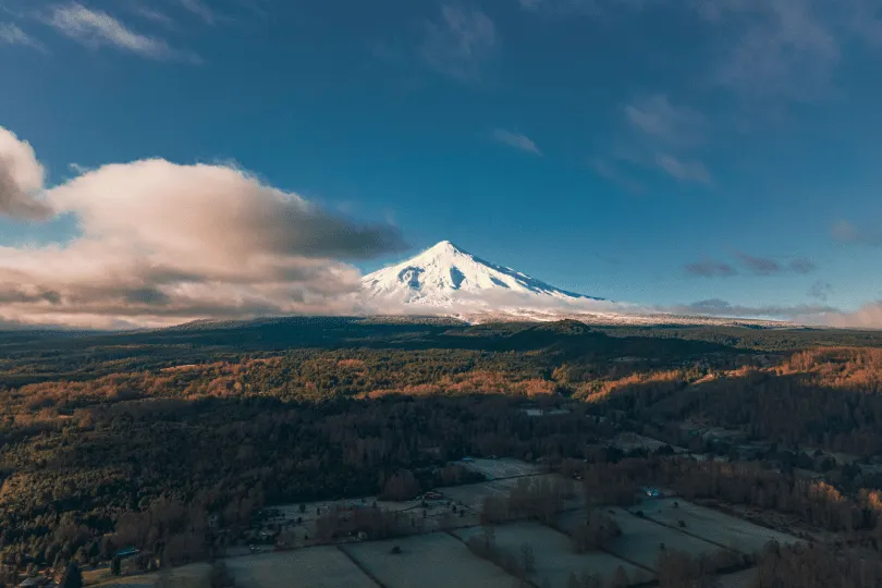 Ein schneebedeckter Vulkan erhebt sich über einer weitläufigen, bewaldeten Landschaft. Tiefe Wolken ziehen an den Berghängen vorbei, während warmes Licht Teile der Wälder und Felder im Vordergrund beleuchtet.