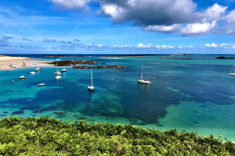 Blick von einer grünen Anhöhe auf eine türkisfarbene Bucht mit mehreren Segelbooten, kleinen Felseninseln und einem Sandstrand unter blauem, wolkigem Himmel.