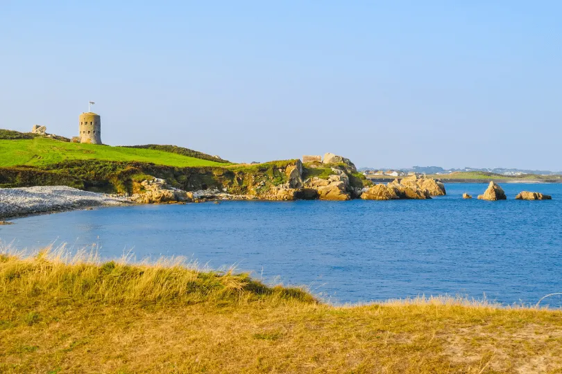 Küstenlandschaft auf Guernsey mit blauem Meer, felsiger Bucht, grünen Wiesen und einem runden historischen Steinturm auf einer Anhöhe.