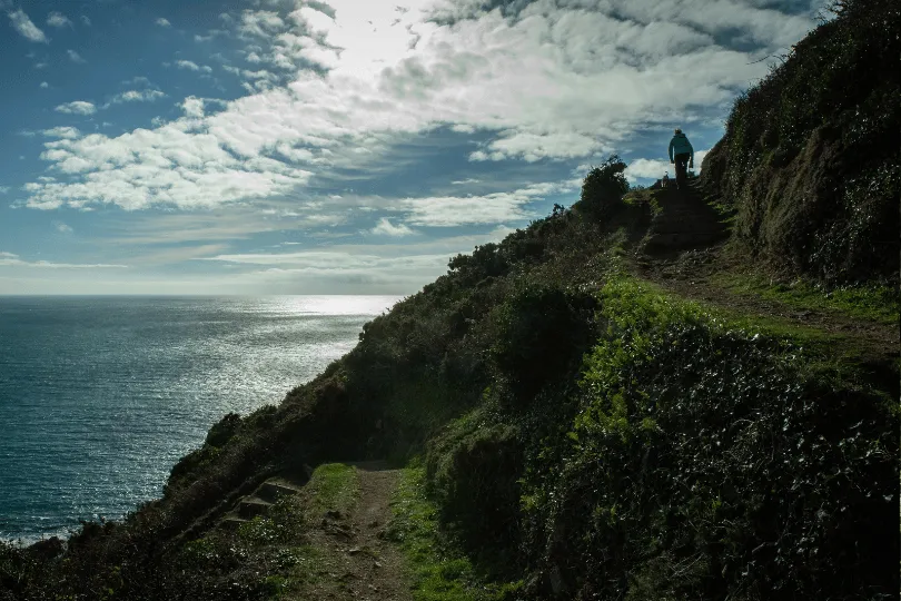 Wanderer auf einem schmalen Küstenpfad entlang der Klippen von Guernsey mit weitem Blick über das Meer unter dramatischem Wolkenhimmel.