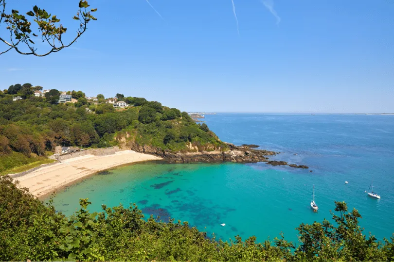 Türkisfarbene Bucht auf Jersey mit Sandstrand, grünen Hügeln, Häusern und Segelbooten unter blauem Himmel.