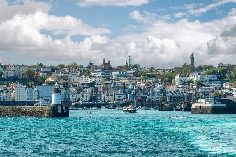 Blick vom Meer auf St. Peter Port auf Guernsey mit Hafen, Leuchtturm und dicht bebauter Küstenstadt unter wolkigem Himmel.