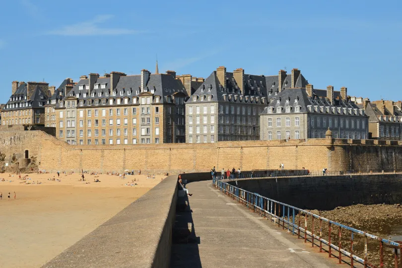 Historische Altstadt von Saint-Malo mit hohen Steinhäusern hinter der Stadtmauer, Sandstrand und einem befestigten Steg am Meer.