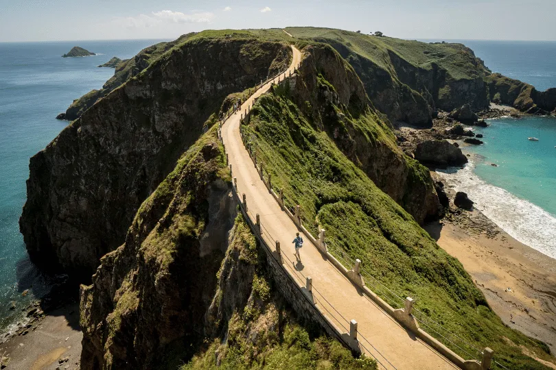 Schmaler Küstenweg auf Sark, der über einen grünen Felsgrat führt, mit einem Radfahrer, steilen Klippen und türkisblauem Meer auf beiden Seiten.