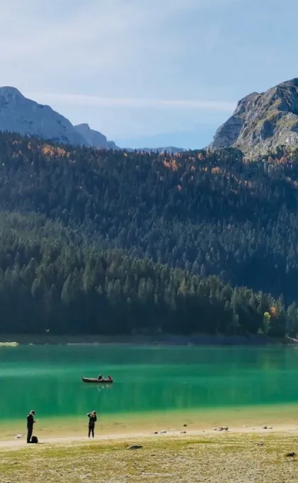 ürkisfarbener Bergsee mit Menschen am Ufer und Boot vor bewaldeten Hängen und felsigen Gipfeln in den Dinarischen Alpen.