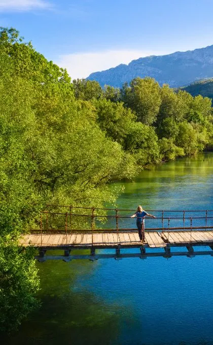 Frau steht auf einer Brücke über einem türkisen Fluss zwischen saftigen Laubbäumen in Montenegro.