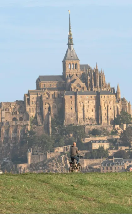 **Alt-Text:** Blick auf den Mont-Saint-Michel in Frankreich mit Radfahrer und Spaziergängern im Vordergrund.