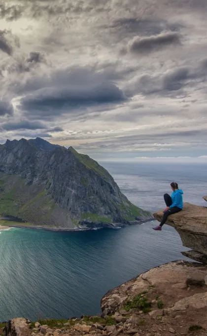 Eine Person in blauer Jacke sitzt auf einem Felsvorsprung hoch über dem Meer und blickt auf eine steile Bergküste unter dramatisch bewölktem Himmel.