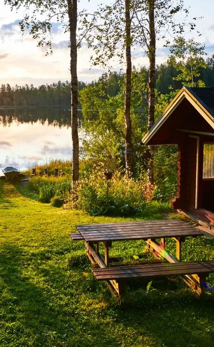 Ein Holztisch und Bänke stehen vor einer Holzhütte direkt am See in Finnland.