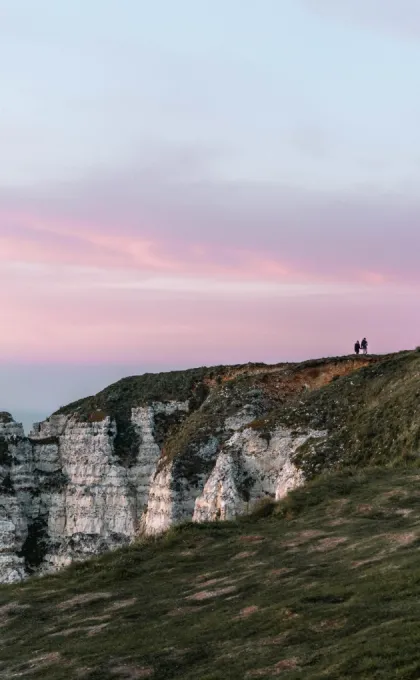 Zwei Personen stehen auf grasbewachsenen Kreidefelsen an der Küste, während der Himmel bei Sonnenuntergang rosa und violett gefärbt ist.