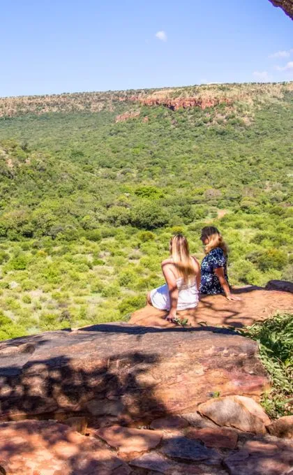 Zwei Frauen sitzen auf einem Felsen mit einer weiten Aussicht über eine Grünfläche in Südafrika.