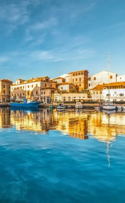 Häuser und Boote in Sardinien strahlen in der Sonne und werden im Wasser reflektiert.