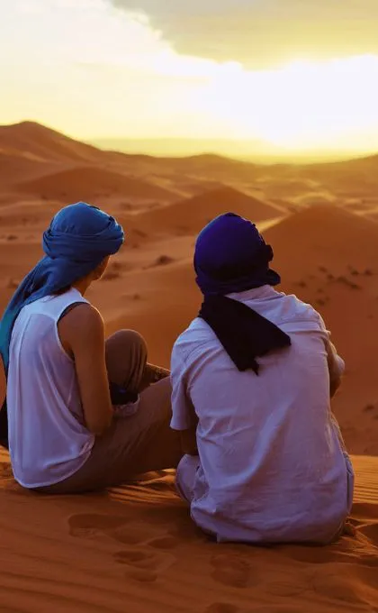 Zwei Reisende in Turbanen sitzen in der Wüste und schauen den Sonneuntergang.