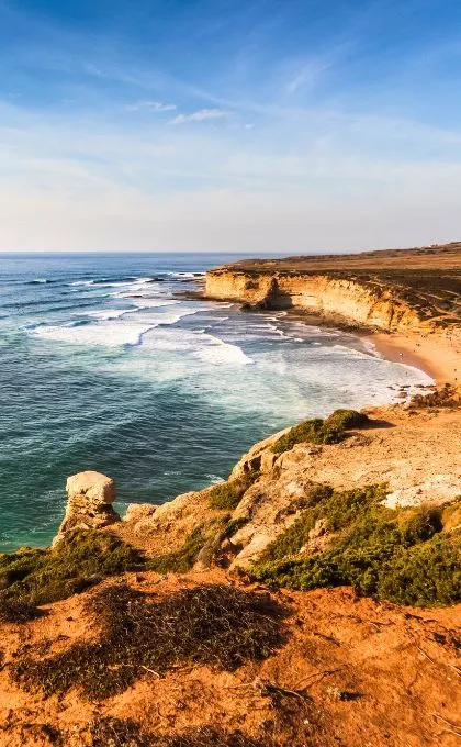 Strand in Portugal mit einer hohen Klippe vor den seichten Wellen des Meers.