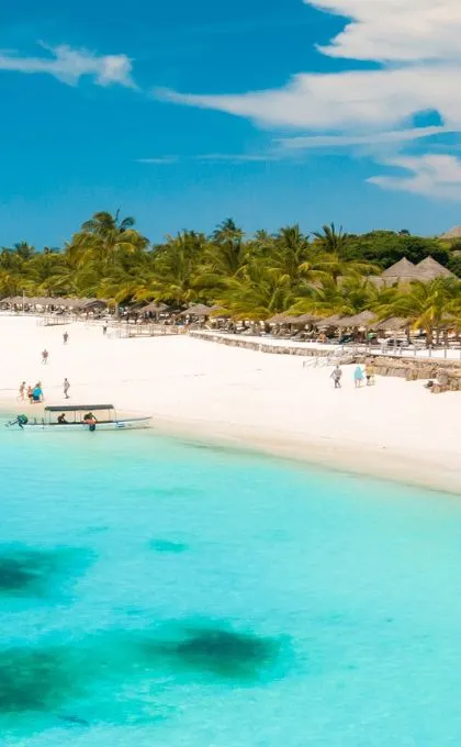 Strahlend weißer Sandstrand am türkisen Wasser in Sansibar. Im Hintergrund stehen einige Lodges zwischen den Palmen.