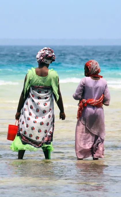 Zweit einheimische Frauen in bunten Gewändern stehen mit den Füßen im Meer in Tansania.