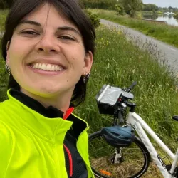 Unsere Fahrradreise-Expertin Johanne steht auf einer Wiese und hält einen Reiseführer in der Hand. Im Hintergrund sind Bäume zu sehen, während sie in die Kamera lächelt.