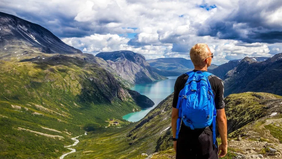 Eine Person mit blauem Rucksack blickt von einem Bergrücken auf ein grünes Tal mit einem gewundenen Fluss.