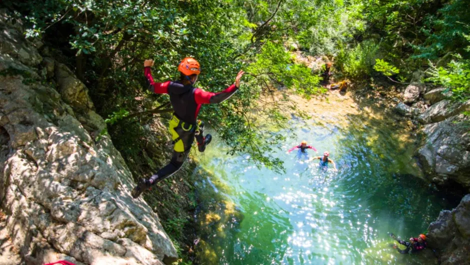 Ein Kind springt beim Canyoning ins Wasser, umgeben von Felsen und Wald. Andere warten bereits im Becken.