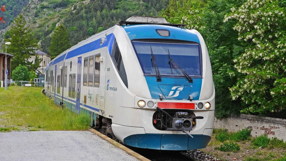 Ein blau-weißer Regionalzug von Trenitalia steht auf einem Gleis neben grünen Bäumen und Hügeln in ländlicher Umgebung.