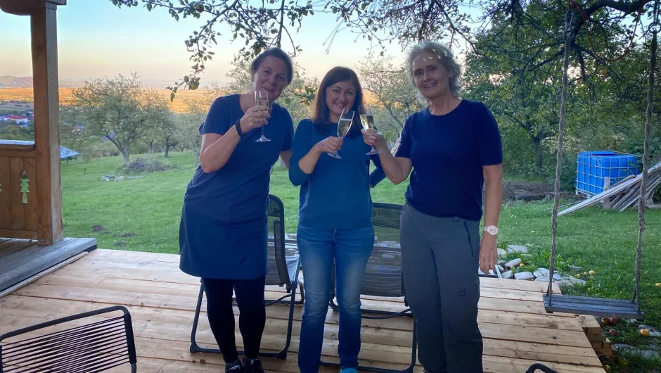 Unsere Reiseexpertin udn Fairaway Chefin Saskia stehen auf einer Holzterrasse unter einem Baum und stoßen mit Sektgläsern an. Im Hintergrund sieht man eine grüne Wiese mit Obstbäumen und eine weite ländliche Landschaft.