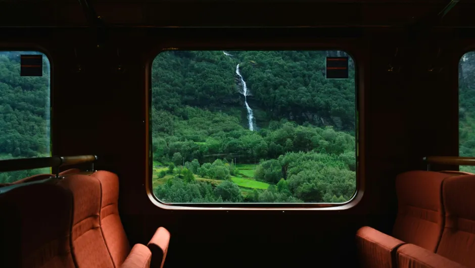 Blick aus einem Zugfenster auf grüne Hügel und einen schmalen Wasserfall; im Vordergrund zwei Sitze im Zugabteil.