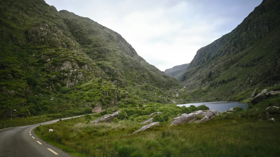 Schmale Landstraße schlängelt sich durch ein grünes Tal zwischen steilen Bergen, vorbei an einem See und vereinzelten Felsen unter bewölktem Himmel.