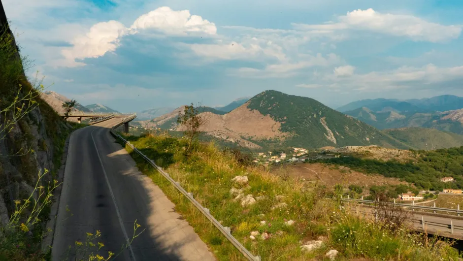 Eine kurvige Bergstraße mit Leitplanke führt entlang eines Hangs, mit Blick auf grüne Hügel, ein kleines Dorf im Tal und entfernte Berge unter einem teils bewölkten Himmel.