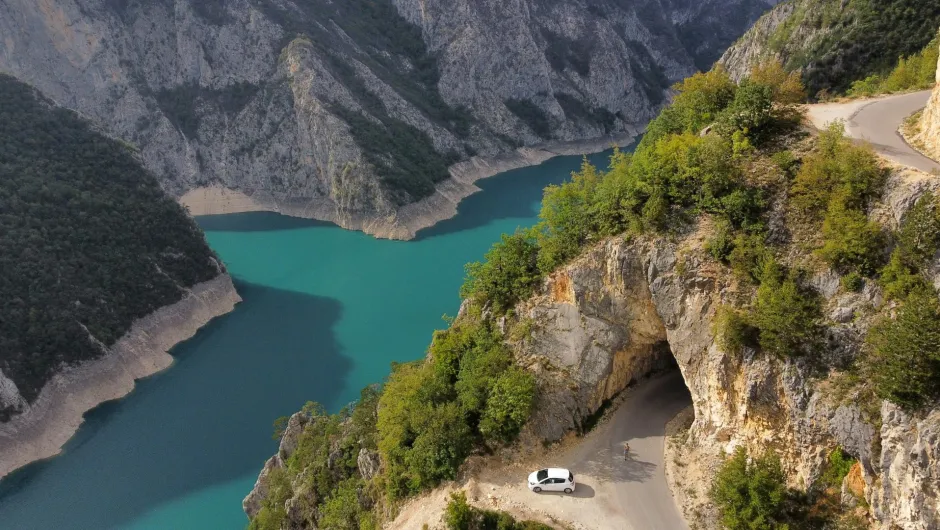 Schmale Bergstraße führt entlang einer steilen Felswand über einem türkisfarbenen See, ein Auto steht nahe eines kleinen Tunnels in der Felswand.