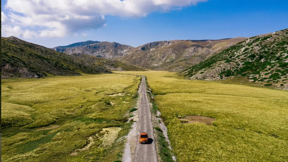 Ein einzelnes Auto fährt auf einer schmalen Straße durch eine weite, offene Landschaft zwischen sanften Hügeln unter blauem Himmel mit Wolken.
