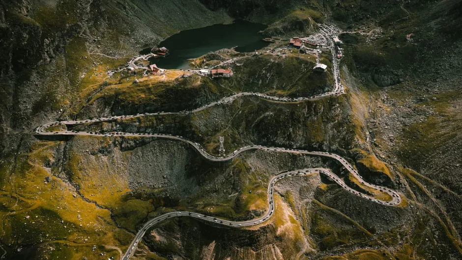 Eine spektakuläre, kurvenreiche Bergstraße schlängelt sich in zahlreichen Serpentinen durch eine felsige Hochgebirgslandschaft. Entlang der Strecke sind viele Autos unterwegs. Oben am Berg liegt ein kleiner See, umgeben von Gebäuden, Parkplätzen und Besuchern.