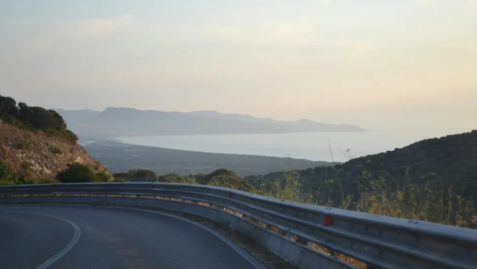 Kurvige Küstenstraße mit Leitplanke führt durch eine hügelige Landschaft mit Blick auf das Meer und entfernte Berge bei warmem Licht.