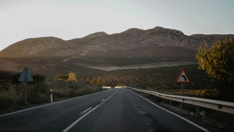 Eine Landstraße führt durch eine bergige Landschaft mit Olivenbäumen; rechts steht ein Kurvenschild, warmes Abendlicht.