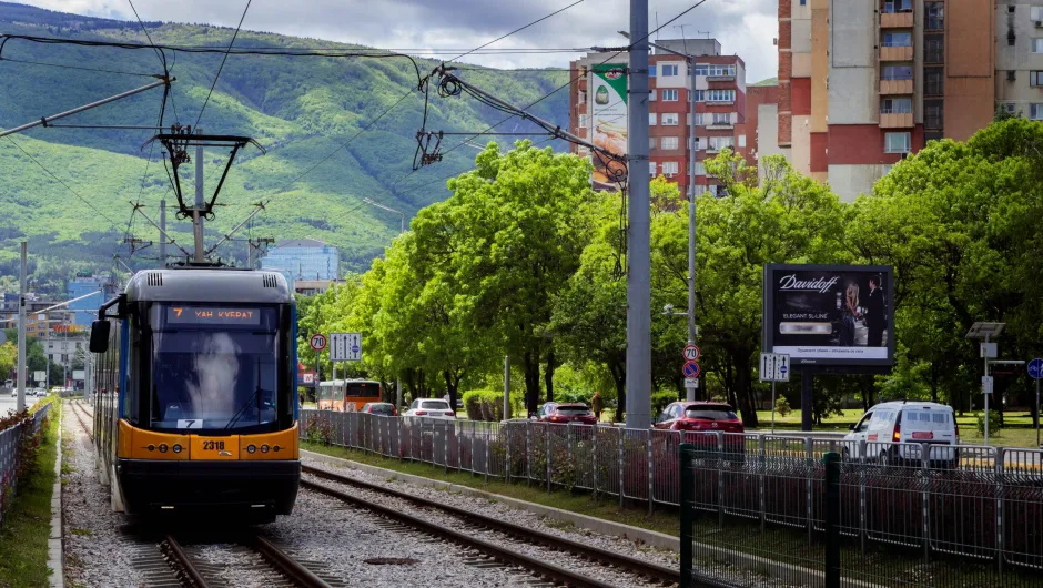 Eine moderne gelb-schwarze Straßenbahn der Linie 7 fährt auf einer begrünten Trasse durch eine Stadtstraße in Sofia. Oberleitungen spannen sich über die Gleise. Links verläuft eine Fahrbahn mit Autos, rechts stehen Wohnblöcke und eine Werbetafel. Im Hintergrund erheben sich grüne bewaldete Hügel oder Berge, die der Szene ein landschaftliches Panorama geben.