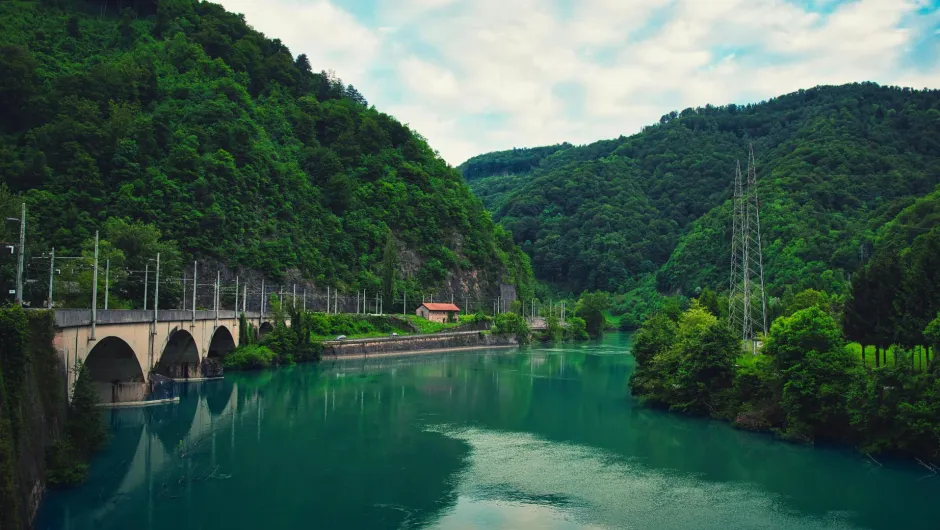 Bahnstrecke auf einer Steinbrücke entlang eines türkisfarbenen Flusses in einem bewaldeten Tal mit grünen Bergen, Strommasten und einem kleinen Haus am Ufer.