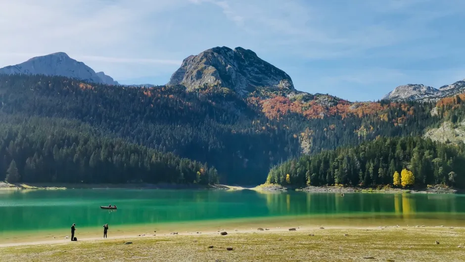 ürkisfarbener Bergsee mit Menschen am Ufer und Boot vor bewaldeten Hängen und felsigen Gipfeln in den Dinarischen Alpen.